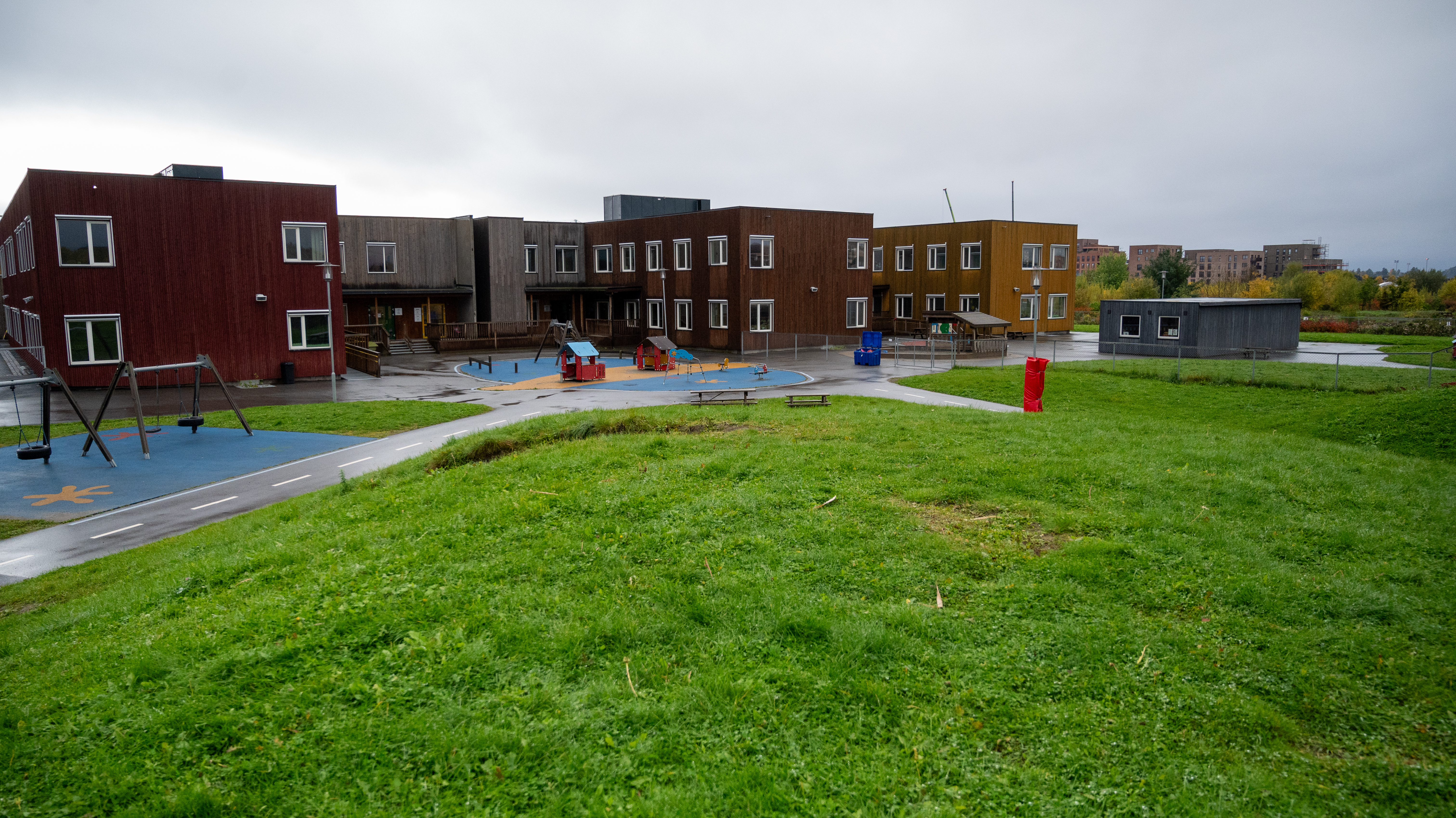 En stor barnehagebygning i flere farger, med to etasjer, står på en åpen gresslette. Foran bygningen er det en lekeplass med husker og små lekehus.