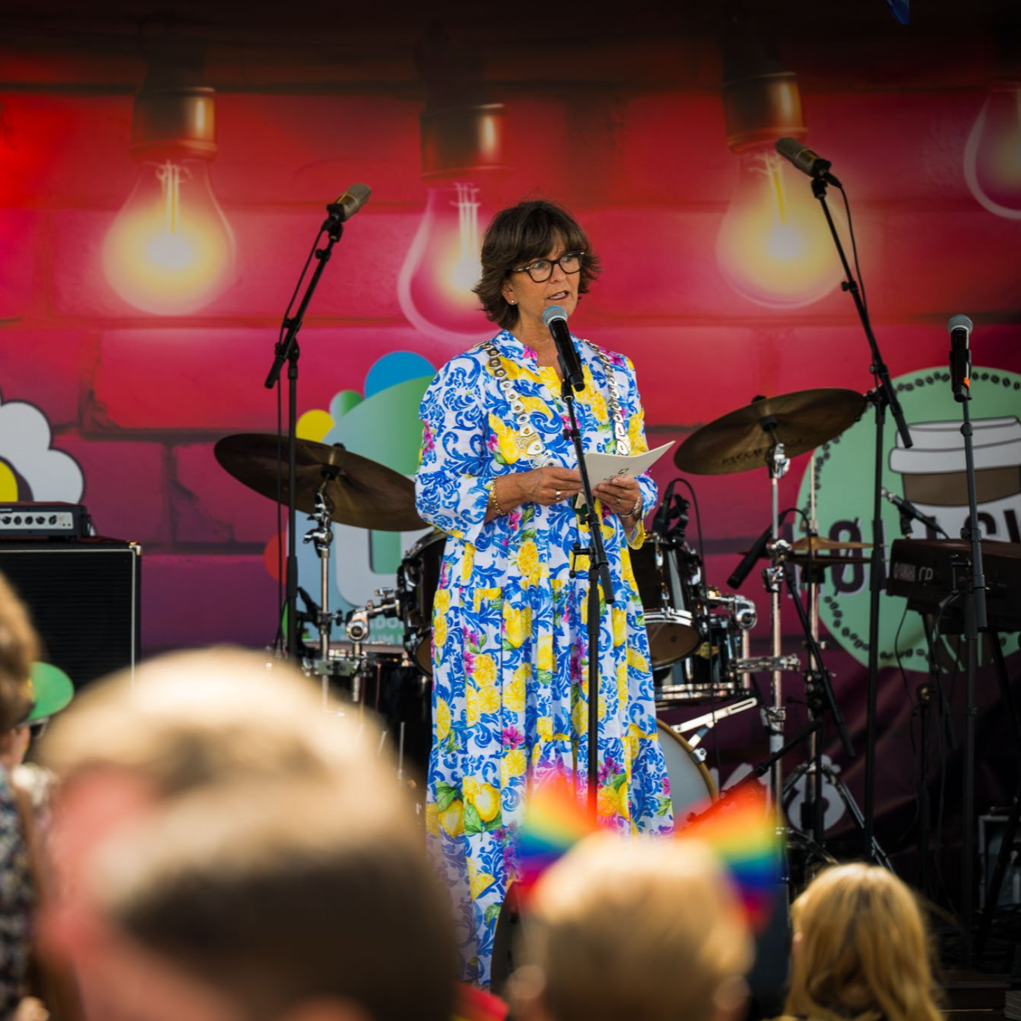 Ordfører Lisbeth Hammer Krog i Bærum står på scenen og holder tale under Sandvika pride 2025, foran et publikum med regnbueflagg og fargerik scenedekor.